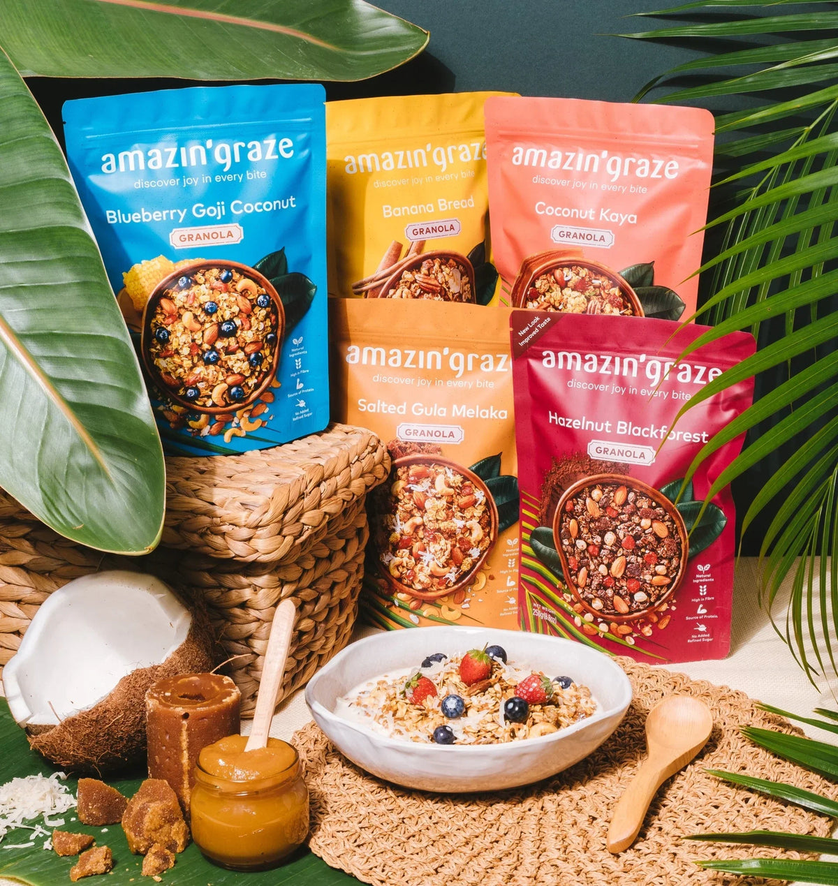 Five Amazin'graze granola pouches in assorted flavors arranged with coconut, honey jar, and a bowl of granola topped with fresh berries.