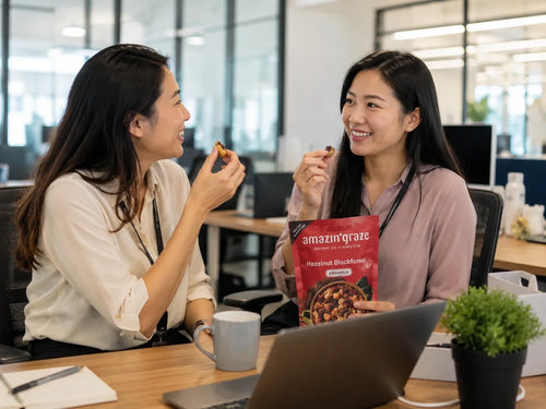 Two women eating Amazin' Graze granola at an office desk while using a laptop.