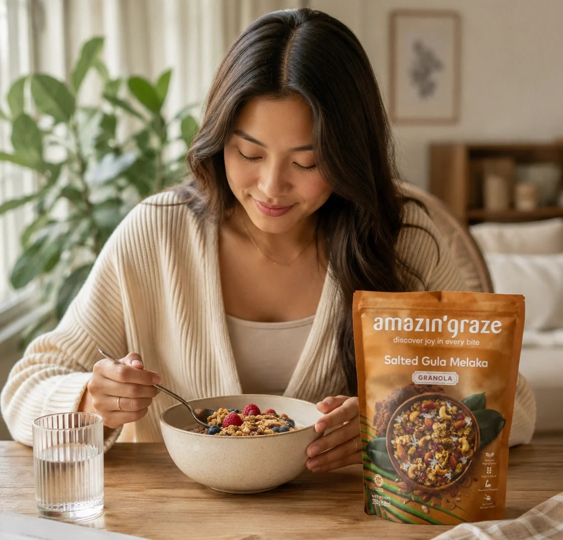 Woman eating Amazin'Graze Salted Gula Melaka granola from a bowl with the product package visible on the table.